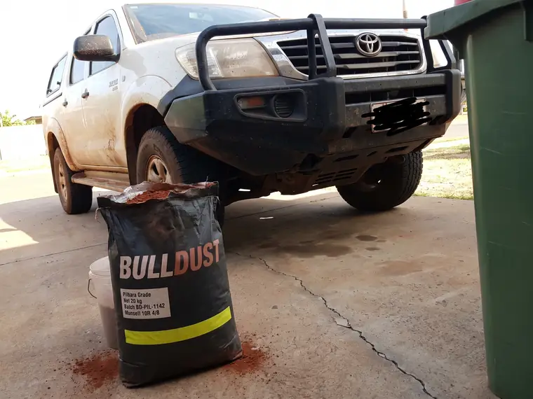 Customer photo of a dusty HiLux with a bull bar parked in a carport, an opened 20 kg Bulldust bag of Pilbara Grade stood on a bucket in front of it. The number plate has been scribbled out.